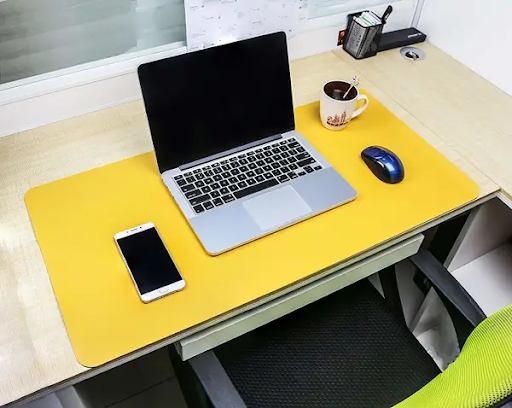 Photo of a large, mostly empty desk. In the foreground, on a yellow mat, a phone, laptop, coffee cup, and mouse are arranged neatly, with plenty of space between them.